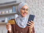Woman in hijab smiling and happy holding smartphone and gold bank credit card